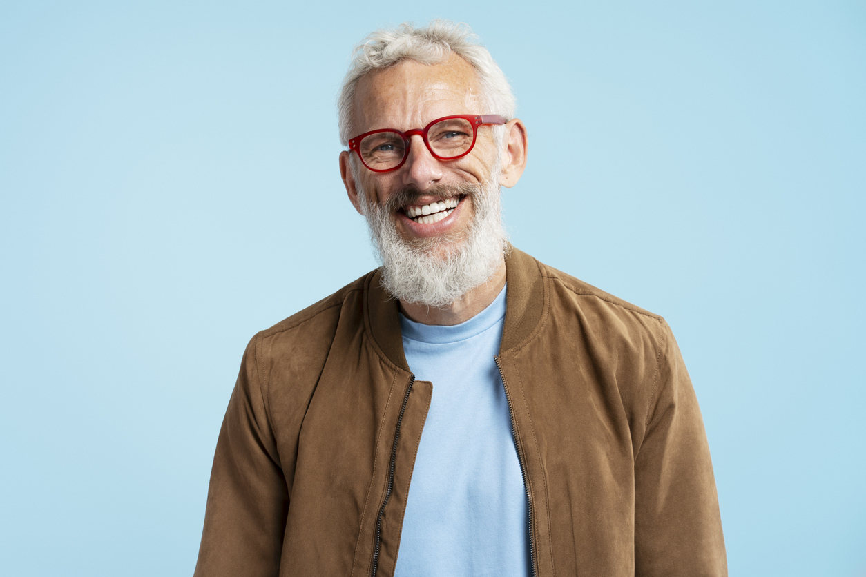 Mature man with dental implants, stylish gray hair wearing red hipster glasses looking at camera isolated on blue background.